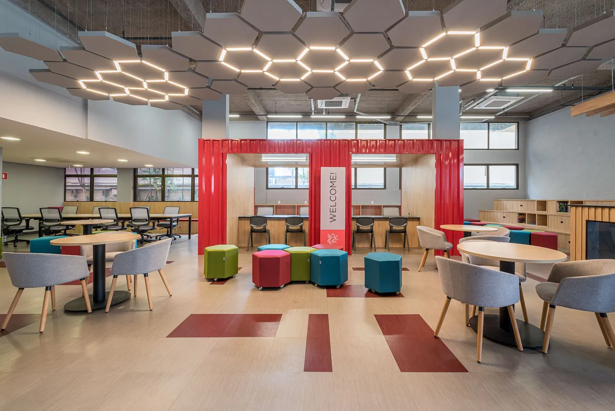Espaço de convivência com luminárias hexagonais no teto, contêiner vermelho de boas-vindas, mesas redondas e pufes coloridos.